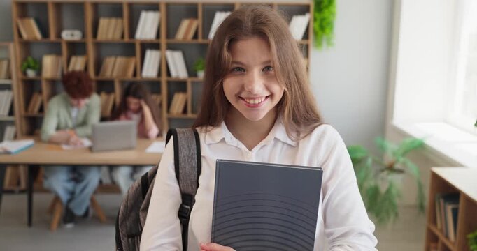 Student girl portrait with smile holding book. Cheerful teenager with backpack in classroom library, ideal for school marketing and education blogs. Clear school learning and success concept.