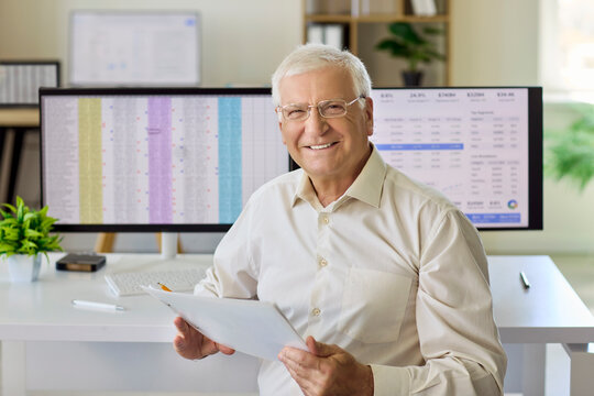 Senior man accountant analyst in office. Smiling professional reviews finance report and spreadsheet on computer screens, signaling analysis and reliability. Clear financial concept shown here.