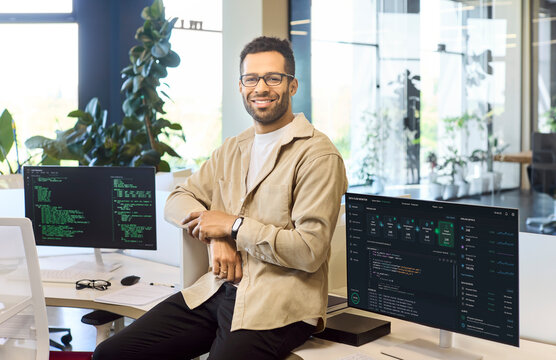 Programmer developer man coding software computer monitor. Modern office workspace features dual monitors with code, clean desk, and plants. Concept shows professional programming and technology.