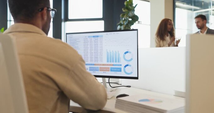 Accountant analyst in office reviewing computer data. Screen shows charts graphs and a financial report as coworkers talk nearby in a bright workspace. Simple concept of finance analysis.