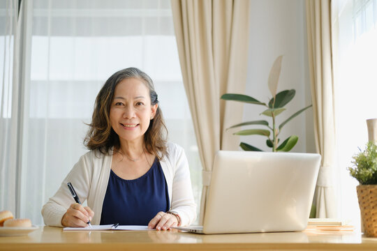 Mature woman smiling while writing notes at a desk with a laptop in a bright home office.