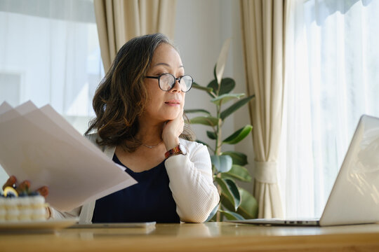 Mature woman wearing glasses and reading documents while working from home at a laptop in a bright office space.