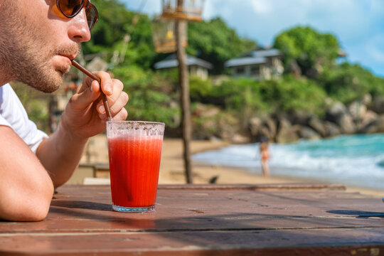 Man tourist in sunglasses drinking refreshing red watermelon drink in glass with straw on wooden table in seaside beach cafe, Samui island, Thailand. Male sipping cold tropical fruit smoothie