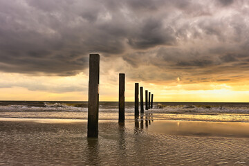 Obraz premium A warm sunset at Zandvoort with dramatic clouds, gentle waves, and wooden posts reflecting light on the wet sand for atmospheric designs.