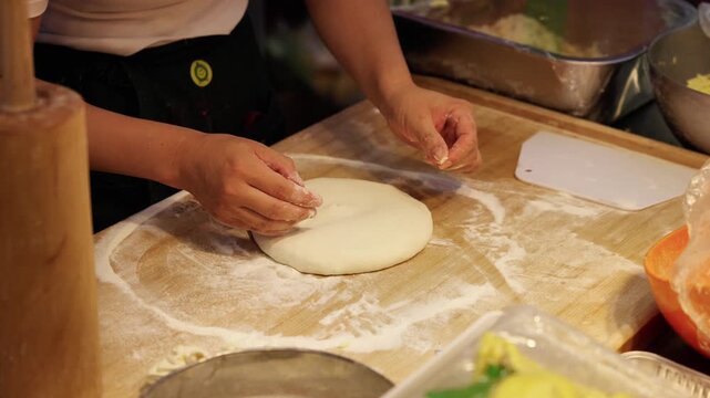 An adult person prepares fresh raw dough on a floured wooden board in a kitchen workspace for food making.