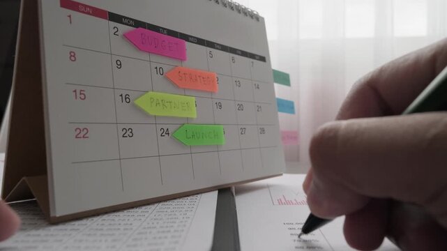 Business person planning timeline on desktop calendar with colorful sticky notes for budget, strategy, partner, and launch, while analyzing financial data and writing on corporate document.
