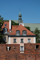 Obraz premium View of vintage European building with tiled rooftop and dormer windows. Red brick fortress wall stands in the foreground. Ancient church tower rises against clear blue sky in Warsaw.