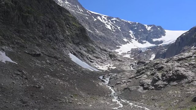 Rocky mountain basin near Sustenpass, Switzerland, with melting snowfields feeding a narrow creek under a clear blue summer sky.