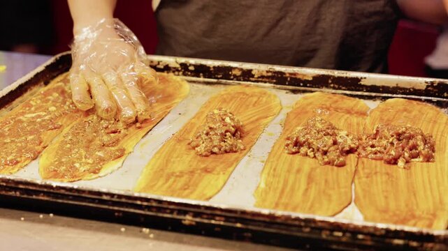 A person prepares traditional tamales, placing filling on corn husks arranged on a metal baking tray.
