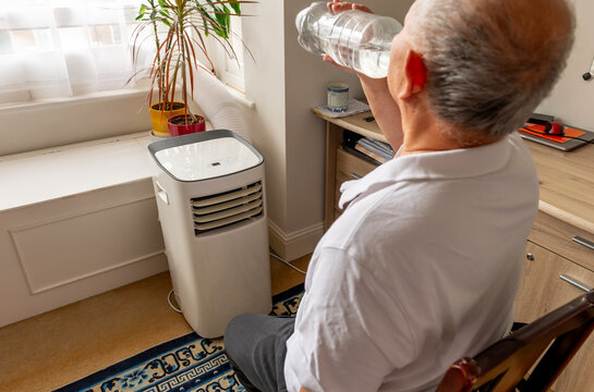 An elderly man cooling down in front of a portable air conditioner and drinking water during a Summer heat wave.