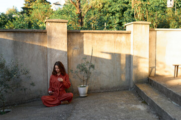 Fototapeta premium A woman in a red outfit sits cross legged on a sunlit courtyard, leaning against a weathered concrete wall, quietly contemplating solitude and inner calm
