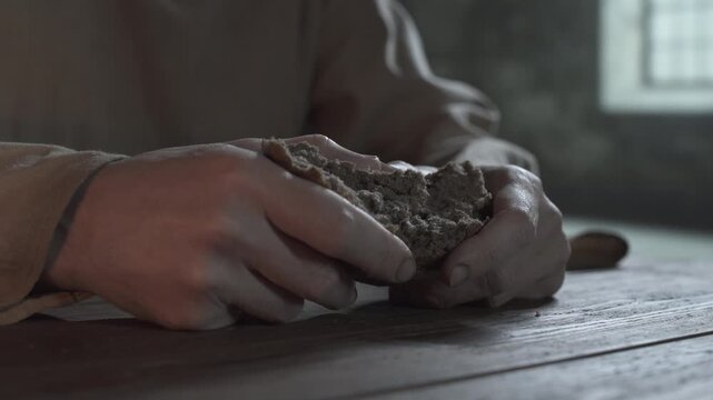Close up of medieval prisoner hands tearing a large loaf chunk of bread on a rustic wooden table inside a gloomy cell