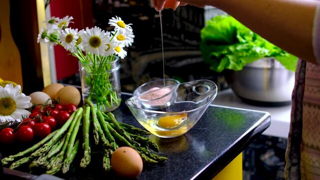 Female hand removing eggshell from raw eggs in glass bowl after cracking fresh eggs with asparagus cherry tomatoes and chamomile flowers for homemade breakfast