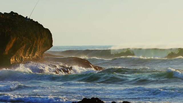 Cinematic 4K slow motion footage of powerful ocean waves crashing against a rugged rocky cliff where a lone fisherman stands. The scene captures the raw beauty of the coastal landscape bathed in the s
