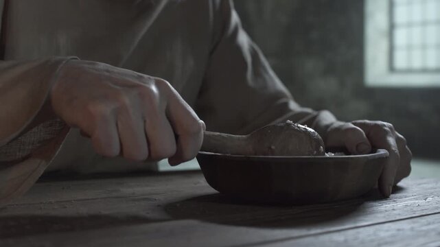 Hands of a medieval prisoner eating porridge with a wooden spoon from a bowl at a rough table inside a gloomy cell