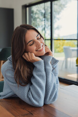 Fototapeta premium Young woman leaning on hands, eyes closed, smiling brightly, feeling peaceful and relaxed at home, enjoying a moment of comfort and tranquility indoors