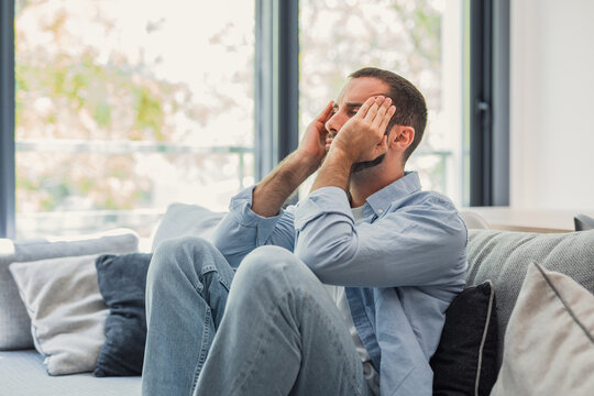 Young man sitting on a sofa at home, pressing his hands to his temples and showing signs of stress, headache and frustration in a bright, casual living room setting.
