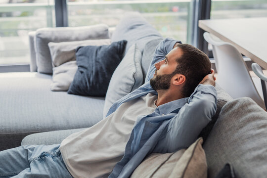 Man with beard finding comfort and peace while relaxing on a modern sofa in his bright apartment, enjoying a moment of quiet contemplation and well-being at home