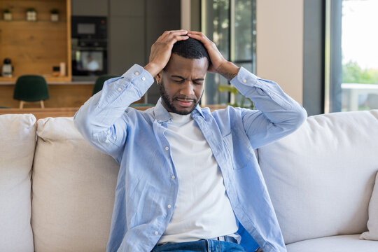 Young man experiencing severe headache and discomfort while sitting on a comfortable sofa at home, feeling frustrated and stressed, needing relief and rest from pain