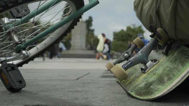 Skateboard and Bicycle Wheel In Extreme Close-Up With Crowd In Backpack Plaza