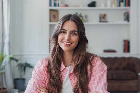 Young woman with long brown hair smiling confidently and looking directly at the camera, wearing a striped pink shirt in a modern home interior with bookshelves in the background