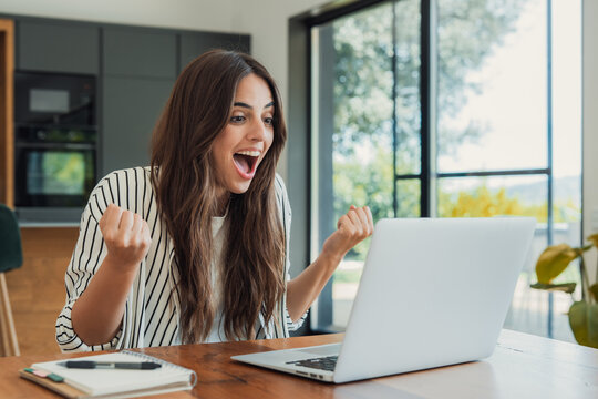 Young woman cheers with enthusiasm at her laptop in a bright home office, celebrating success during remote work or a video call. Natural light, casual outfit, productivity and positive emotions.