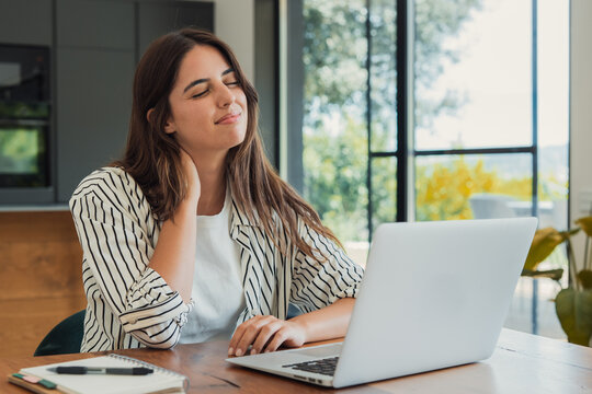 Woman feeling neck pain and stiffness while working from home on a laptop, rubbing her neck, experiencing discomfort from a bad posture and long hours, reflecting professional burnout and stress