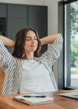A young woman leans back with eyes closed at a wooden desk in a bright home office, taking a peaceful work break next to a laptop and notebook, enjoying calm and focus.