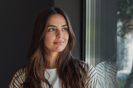 Thoughtful young brunette woman gazes out a window, bathed in soft natural light with a subtle reflection on the glass, conveying calm, introspection and quiet confidence in a casual setting.