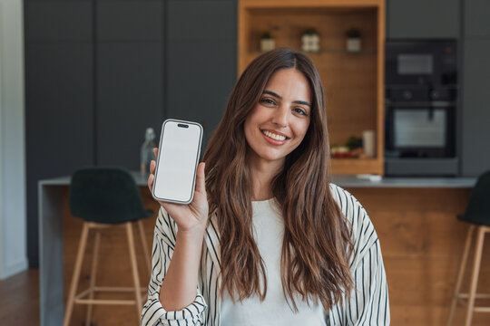 Smiling young woman at home holds up a smartphone with a blank screen in a modern kitchen, conveying happiness, trust and mobile technology for app demos, social media, remote work and lifestyle.