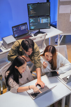 Three young Asian professionals - one man and two women - collaborate closely around a laptop, reviewing programming code together. Multiple monitors displaying code editors glow in the background 