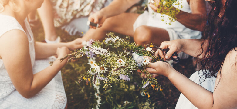 Young women weaving a wildflower wreath before a traditional summer solstice ceremony. Hands creating a floral crown from fresh meadow flowers, celebrating midsummer customs.