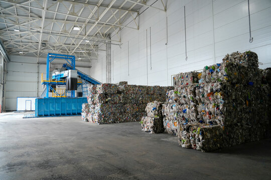 Bales of sorted plastic materials are stacked high inside a spacious recycling plant. Blue sorting machinery with conveyors stands ready to process more recyclables