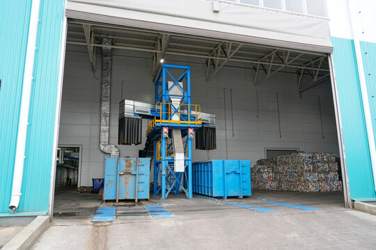 Inside a spacious waste processing facility, blue industrial equipment handles materials, with collection bins and recycled bales awaiting further processing