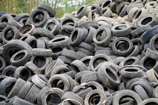 Hundreds of dark, worn vehicle tires are stacked haphazardly outdoors at a waste disposal site under daylight