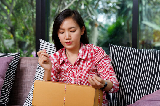 Focused Asian woman tying string on a cardboard parcel box while sitting on sofa. Online seller packing product for customer delivery at home. SME business.