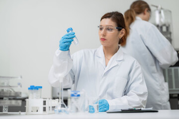 Fototapeta premium Woman in a white lab coat holds a test tube. She wears blue gloves and safety glasses. Lab equipment sits on the workbench. Another scientist works in the background