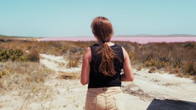 Rear view of young adult woman walks towards a pink lake on a sunny day. She stands on a sandy path surrounded by grass and plants.