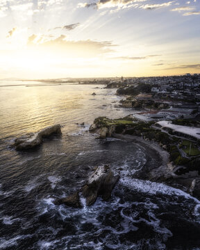 Aerial view of the coastline at Newport Beach featuring rocky outcrops, crashing waves, and coastal architecture under a golden sunset sky in Newport Beach, California, United States.