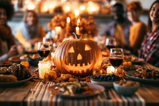 Friends having a cozy autumn dinner party with delicious food and wine on the table, carved pumpkin centerpiece and warm candle lighting