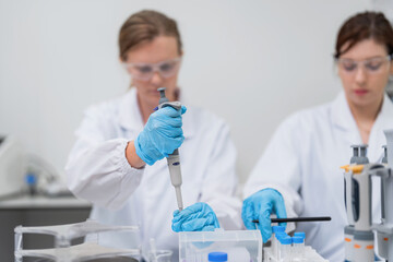Fototapeta premium Two lab technicians wear white coats and blue gloves. One uses a pipette for liquid measurement. The other manages lab equipment and containers. Background includes typical laboratory apparatus