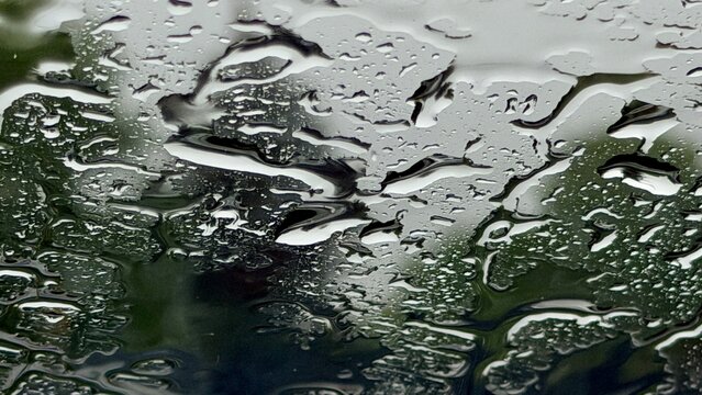Abstract macro view of heavy rain streams on window glass with distorted green trees background. Artistic look of fresh raindrops and wet windowpane texture during stormy summer season weather.