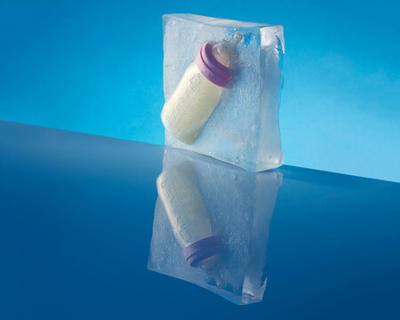 A baby's bottle full of milk is frozen in a block of ice against blue