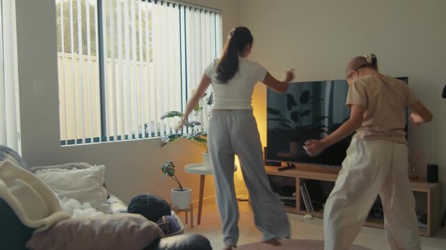 Mother and her daughter cheerfully dance in a living room filled with light from windows. They seem to enjoy their time together while moving.
