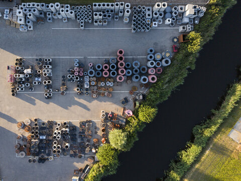 Aerial view of a construction supply yard featuring rows of concrete pipes, rings, and industrial materials arranged on a paved lot beside a canal in Middenmeer, North Holland, Netherlands.