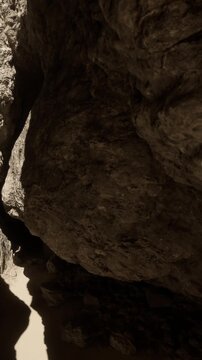 A person navigates a tight space formed by two large rocks. The area is bright from sunlight, highlighting the textures of the stones. The ground is sandy and uneven.