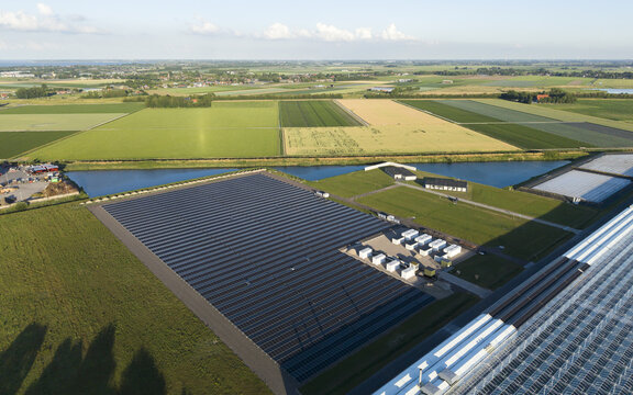 Aerial view of a large solar farm with rows of photovoltaic panels, adjacent to a greenhouse complex and agricultural fields separated by a canal in Middenmeer, North Holland, Netherlands.