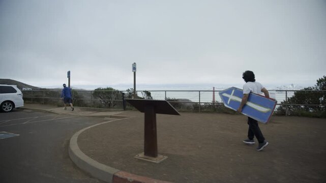 Man Running with Blue Cross Board In Coastal Parking Lot - Wide, Steady Cam Shot