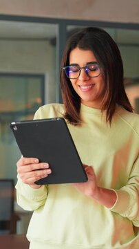 Focused latin young adult professional business woman working on digital device pad gadget. Indian female manager or it specialist holding digital tablet using fintech tab for work in office. Vertical
