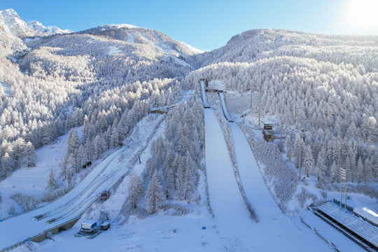 Aerial view of the Stadio Olimpico del Salto ski jumping facility surrounded by snow-covered pine forests and mountains in Pragelato, Piedmont, Italy.